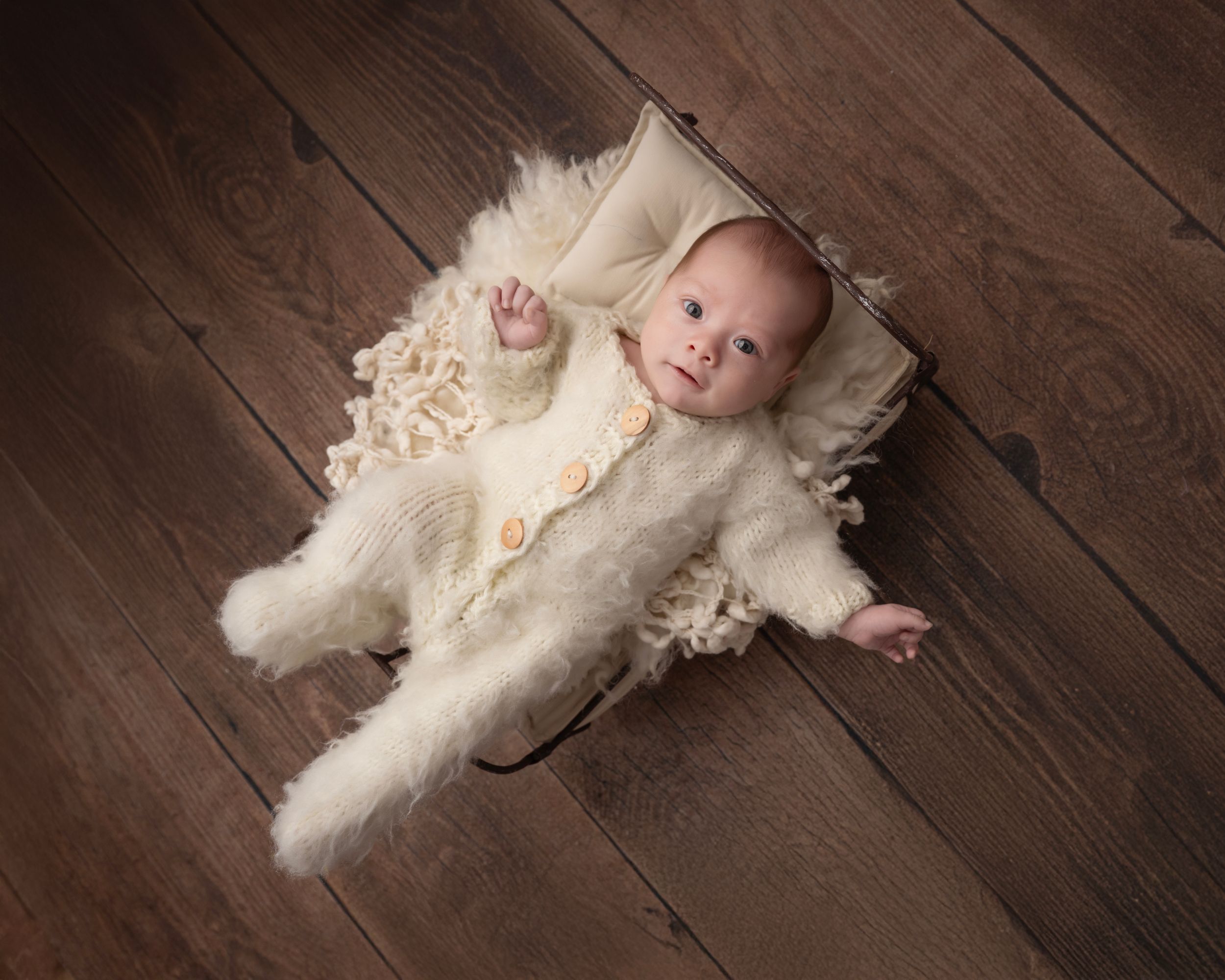 Baby in cream outfit photographed on dark wooden floor with soft lighting.