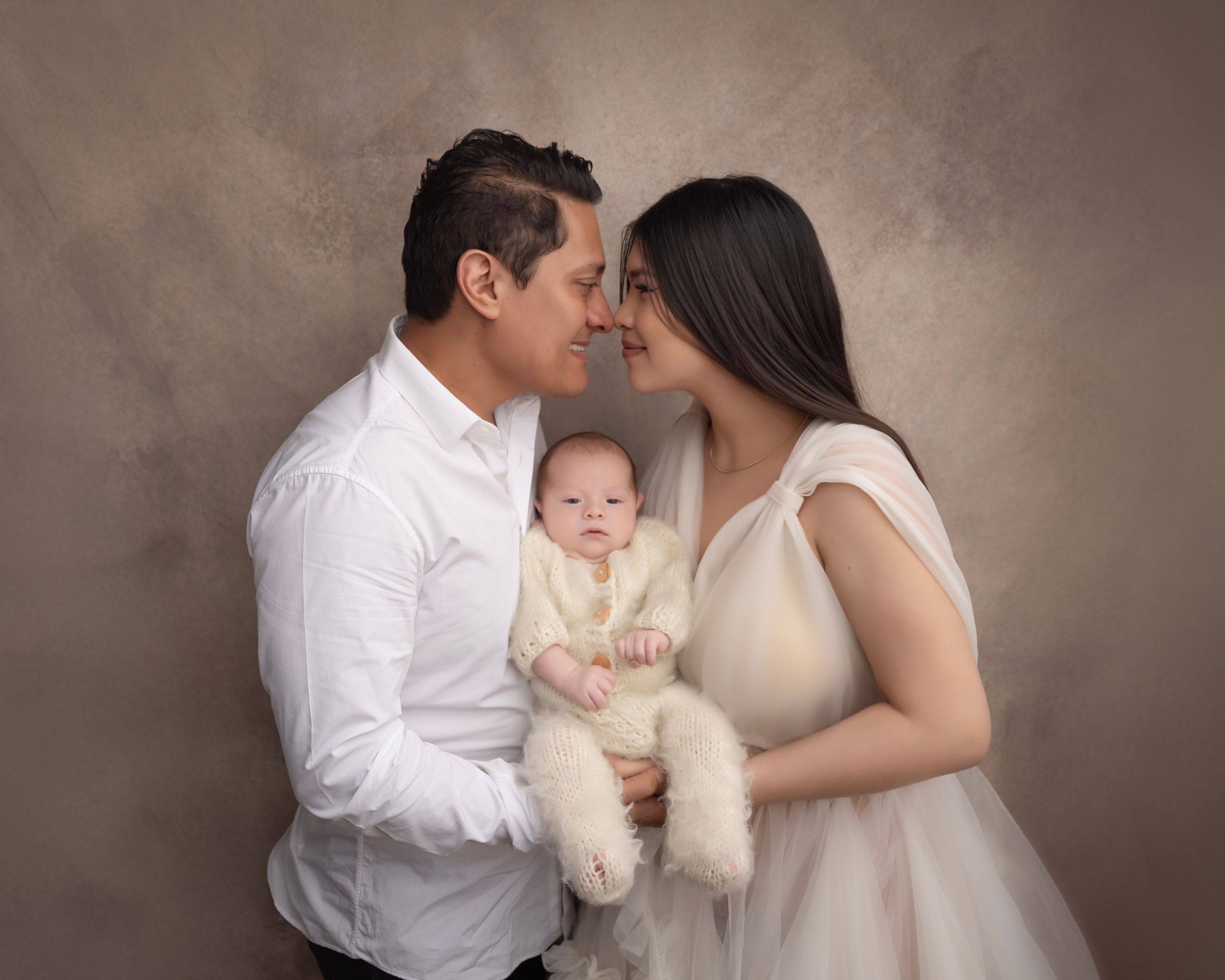 Family portrait in soft brown tones with parents and baby in matching cream outfits.