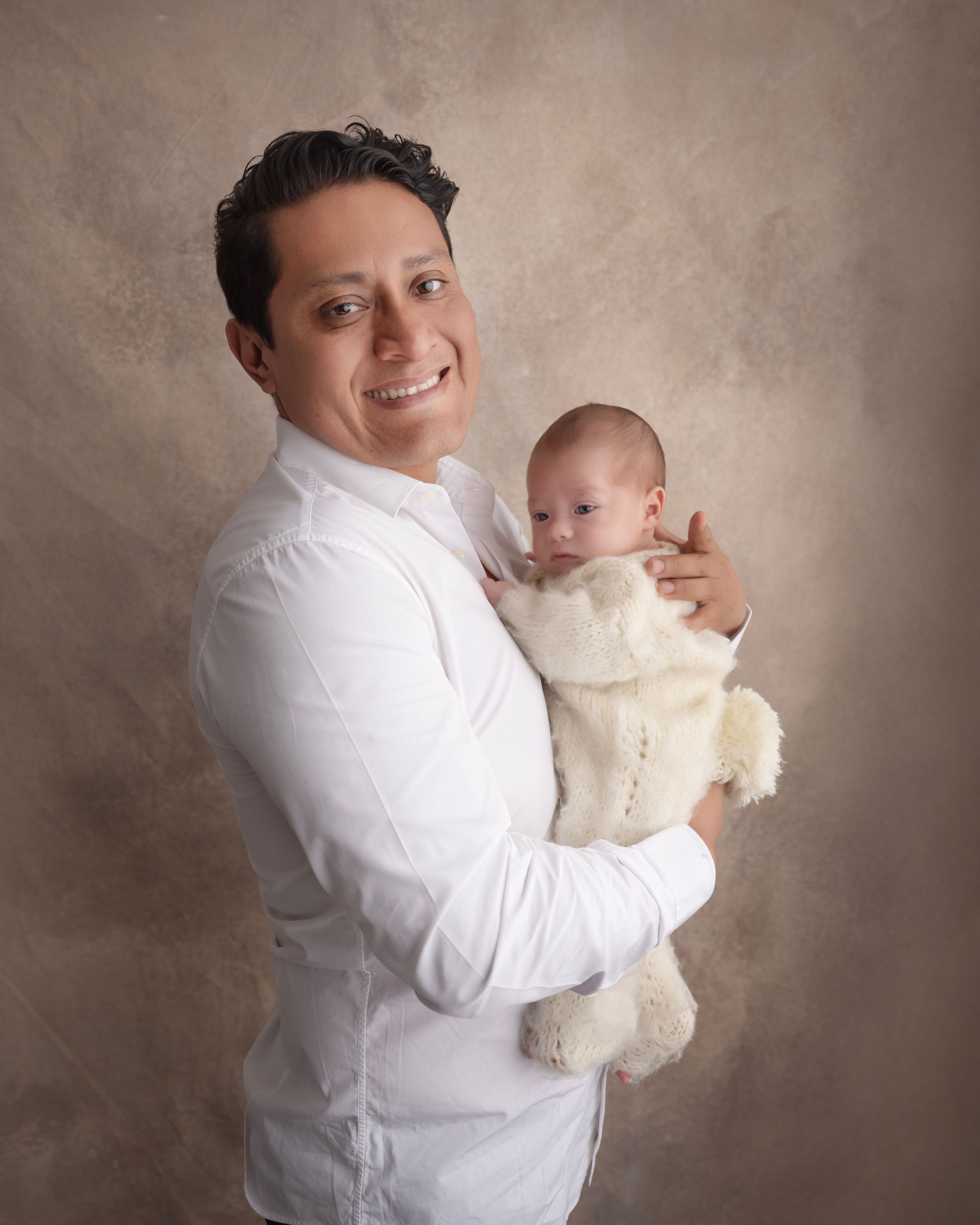 Father in white shirt holding baby against neutral brown studio backdrop.