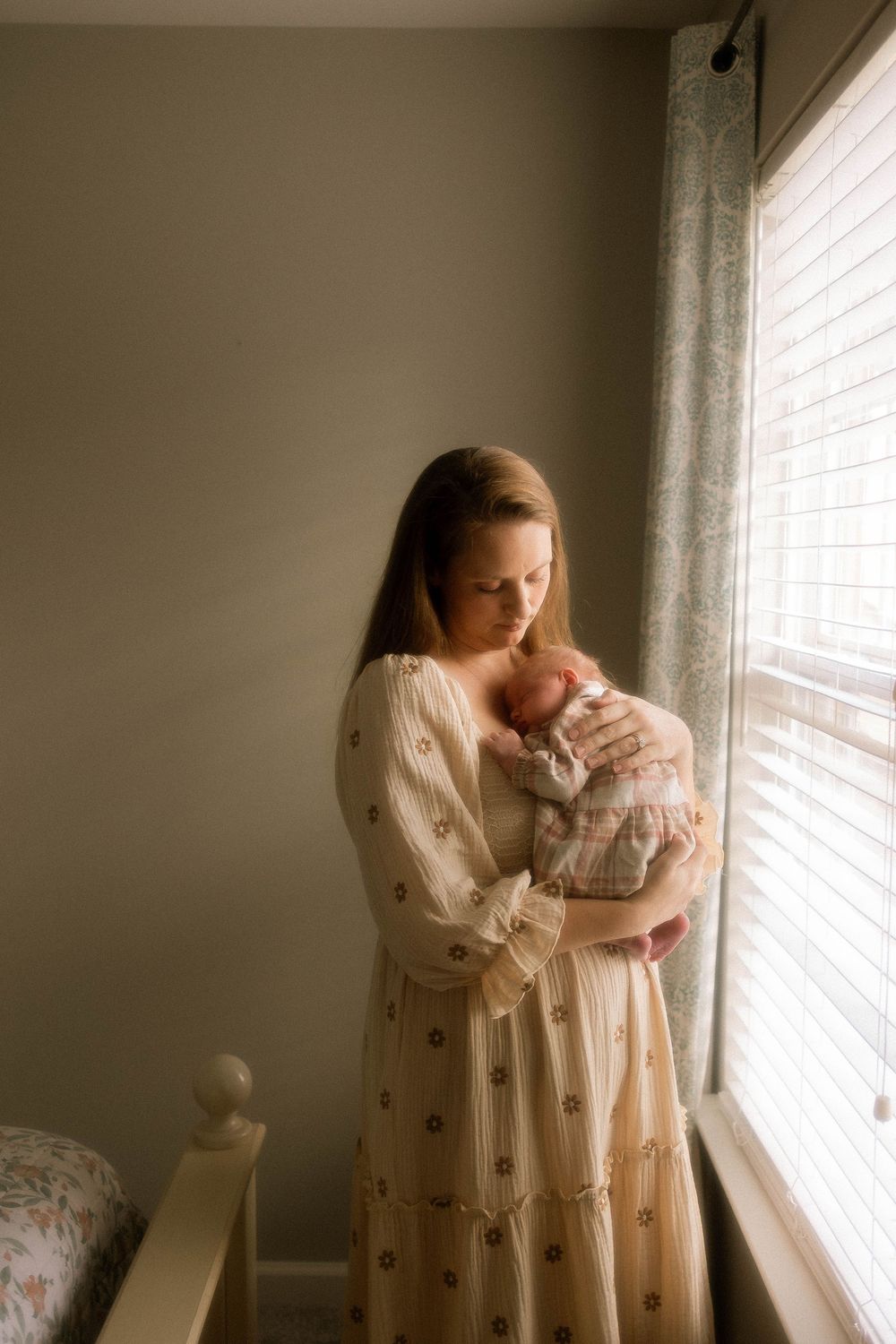 A tender moment by the window as a parent in a floral dress cradles their sleeping newborn in soft natural light.