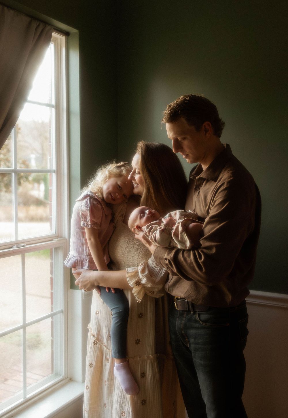 A couple stands in an intimate embrace by a sunlit window in a dimly lit room.