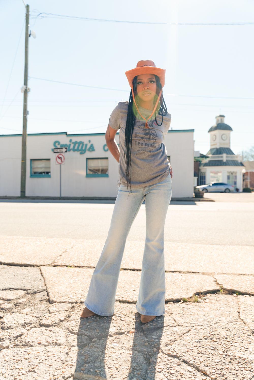 A person in jeans and orange hat stands in a sunny outdoor parking lot near a commercial building.