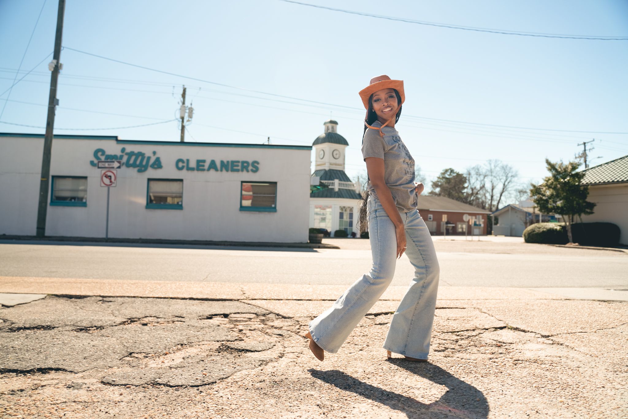 A person in tan overalls and an orange beanie poses playfully in front of a small cleaners storefront on a sunny day.