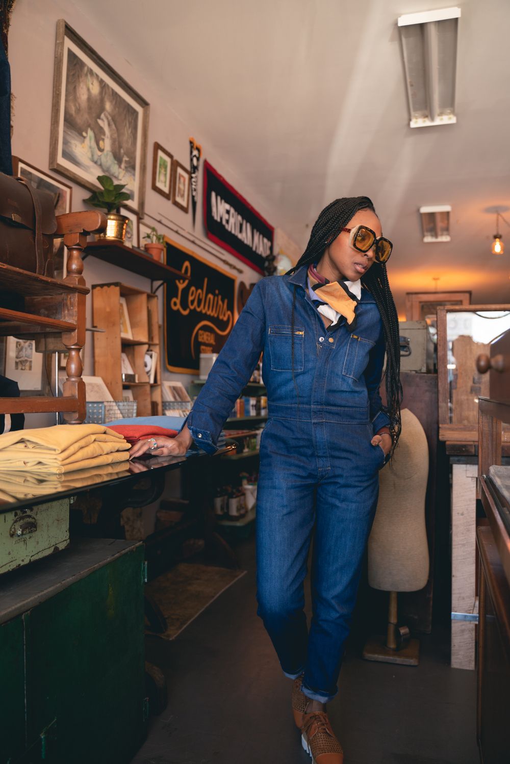 Stylish person in denim overalls and glasses poses casually inside a vintage-style retail shop with rustic decor.