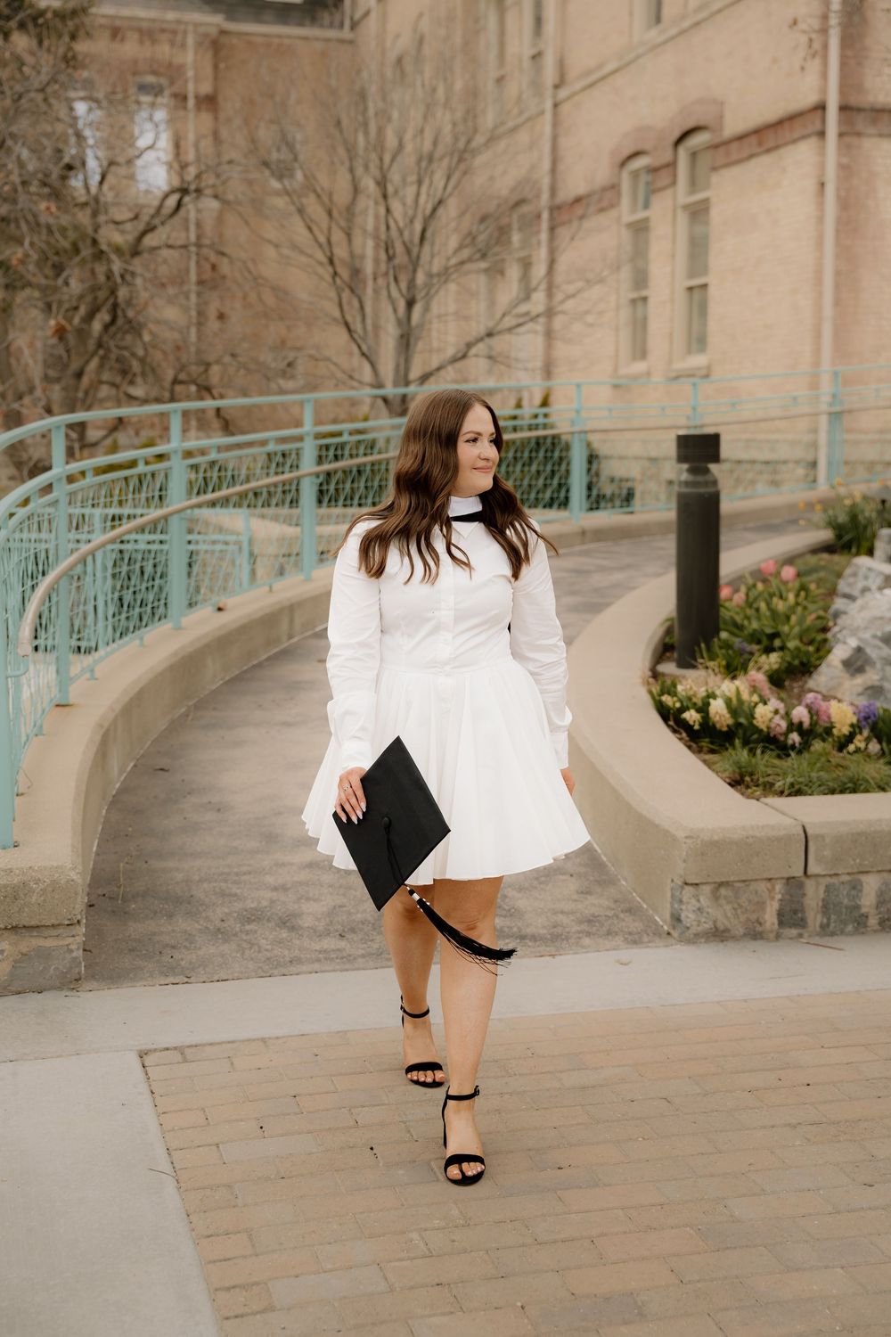 A woman in a knee-length white dress and sandals poses on a curved walkway near a stone building.