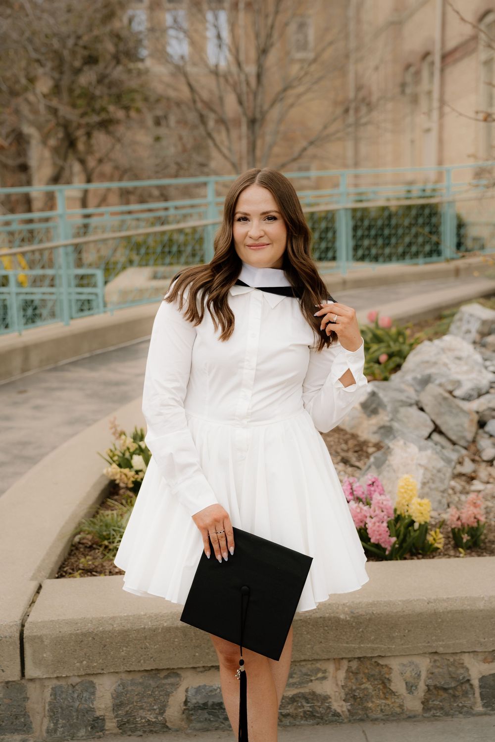 A person in a white long-sleeved dress carries a black portfolio while standing near flower beds and stone walls.
