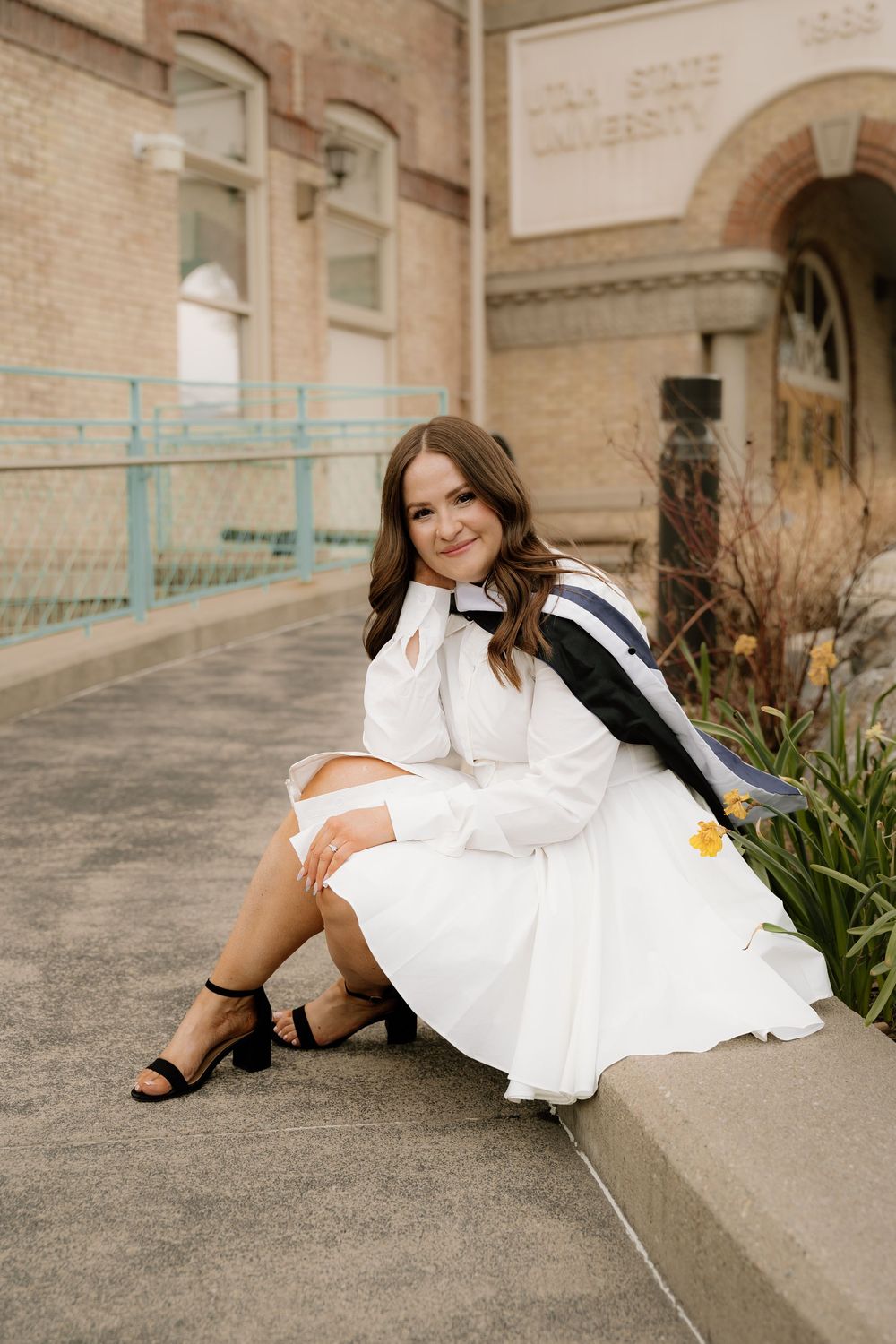 Graduation portrait series with white dress and black heels against brick building background.