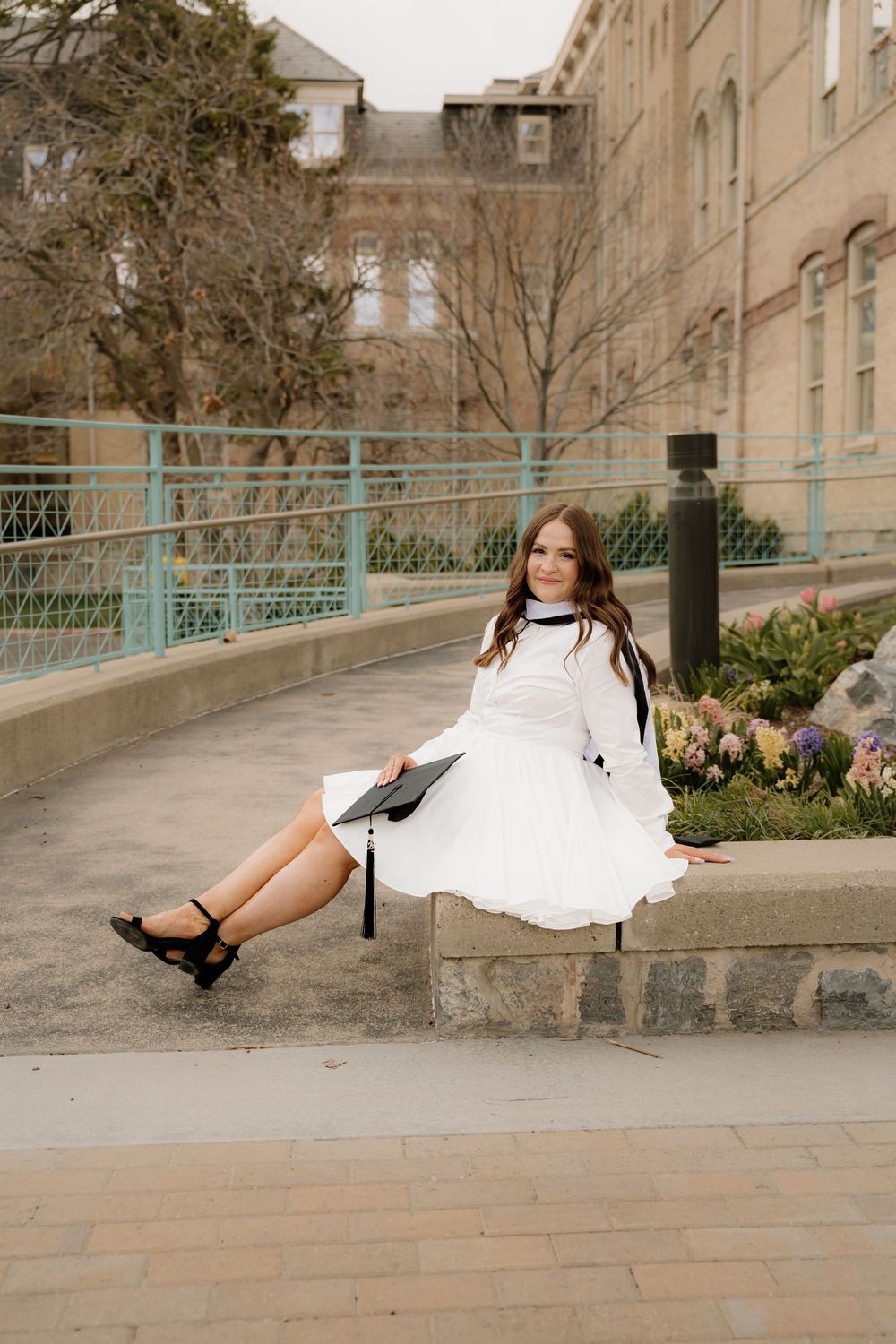 A person in a white dress sits casually on a stone ledge near a vintage building with arched windows and teal railings.