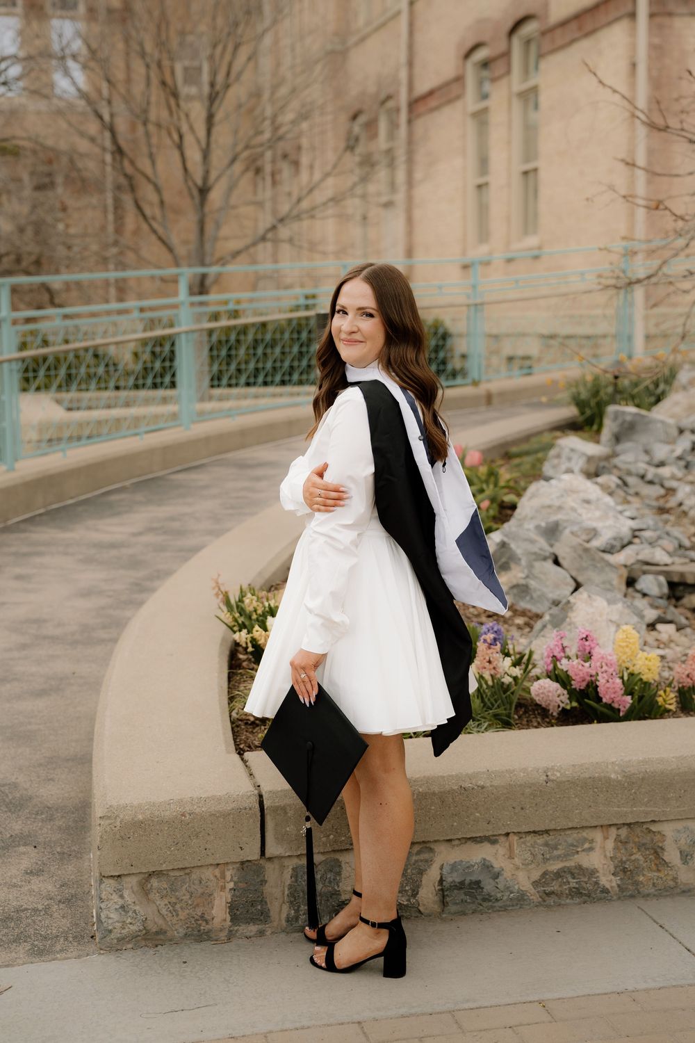 Graduate in white dress and black graduation robe poses outdoors with diploma holder and floral garden backdrop.