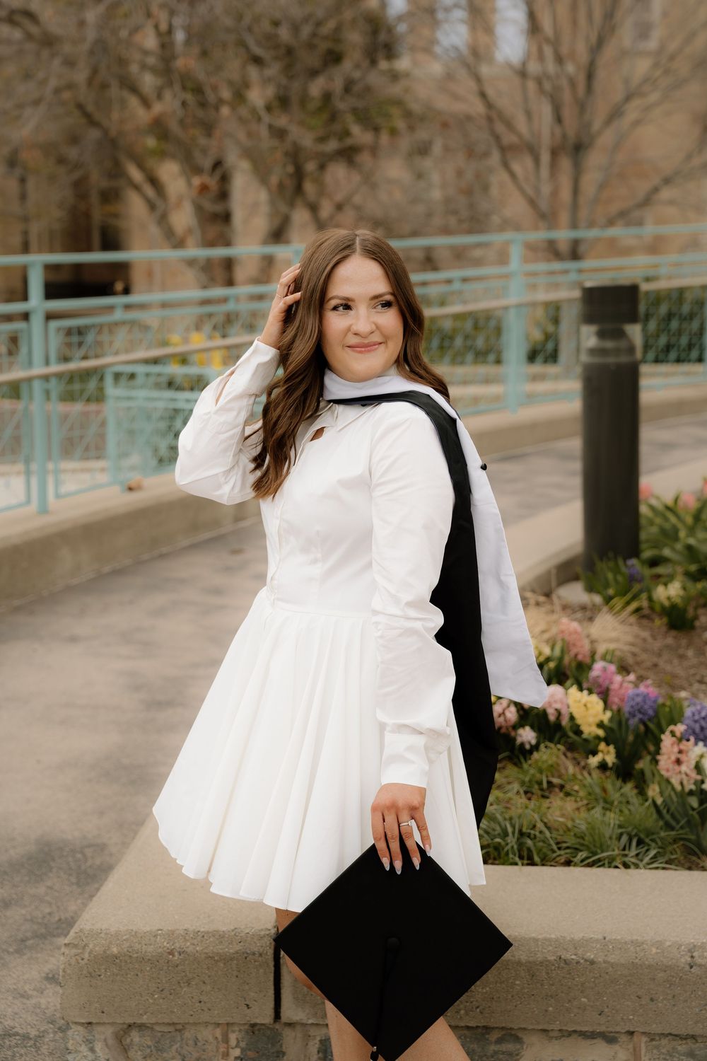 A person in a white dress holds a graduation cap while standing on a pathway with flowers and railings in the background.