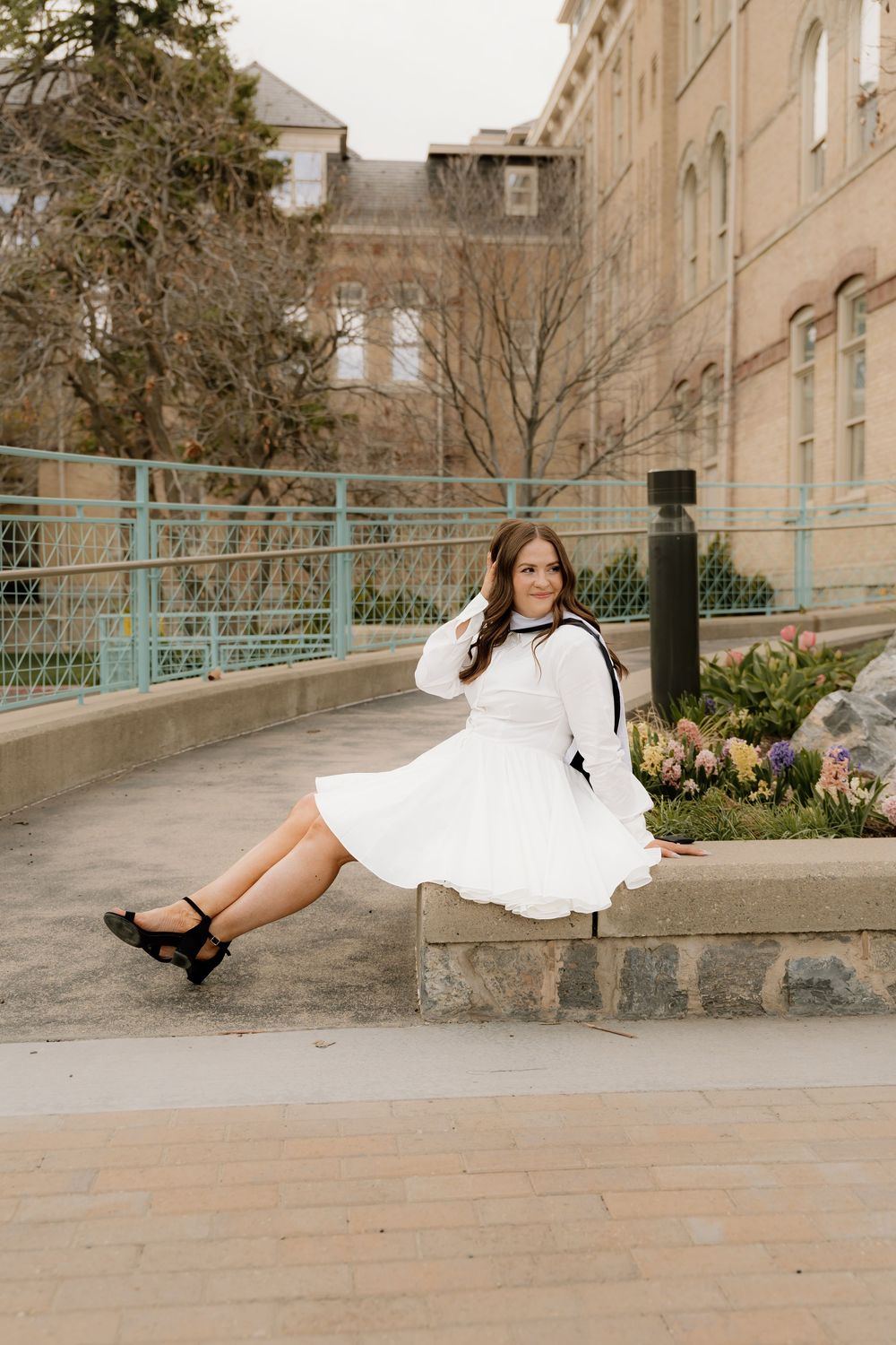 Someone in a white flowy dress and black cardigan sits on a stone wall near flowering tulips on a spring day.