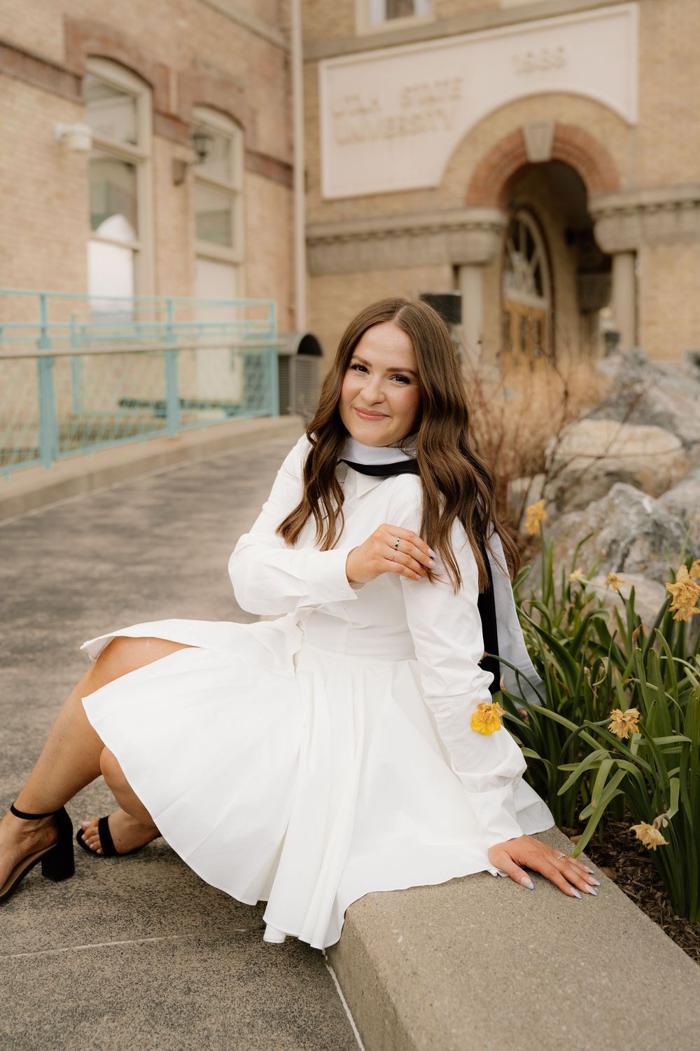 A person in a flowing white dress and sweater sits on a concrete ledge near blooming daffodils outside a historic building.