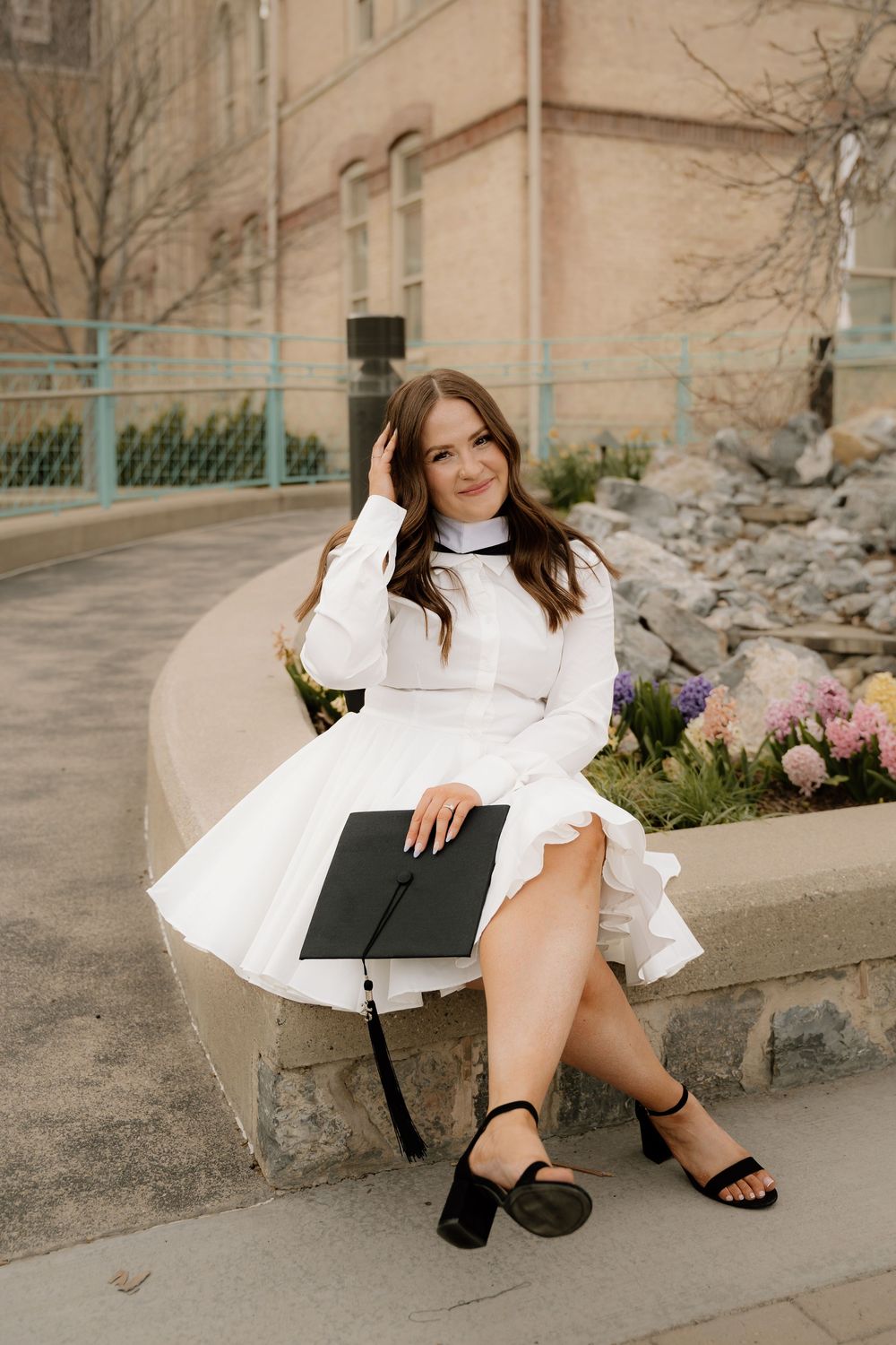 Graduate in a flowing white dress holding a diploma sits on a curved wall with flowers beside a building.