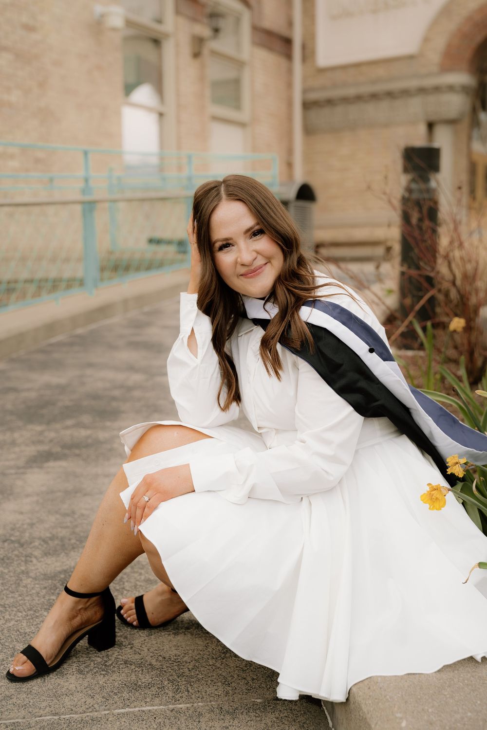Graduate in white dress and black heels sits gracefully on steps during outdoor portrait session.