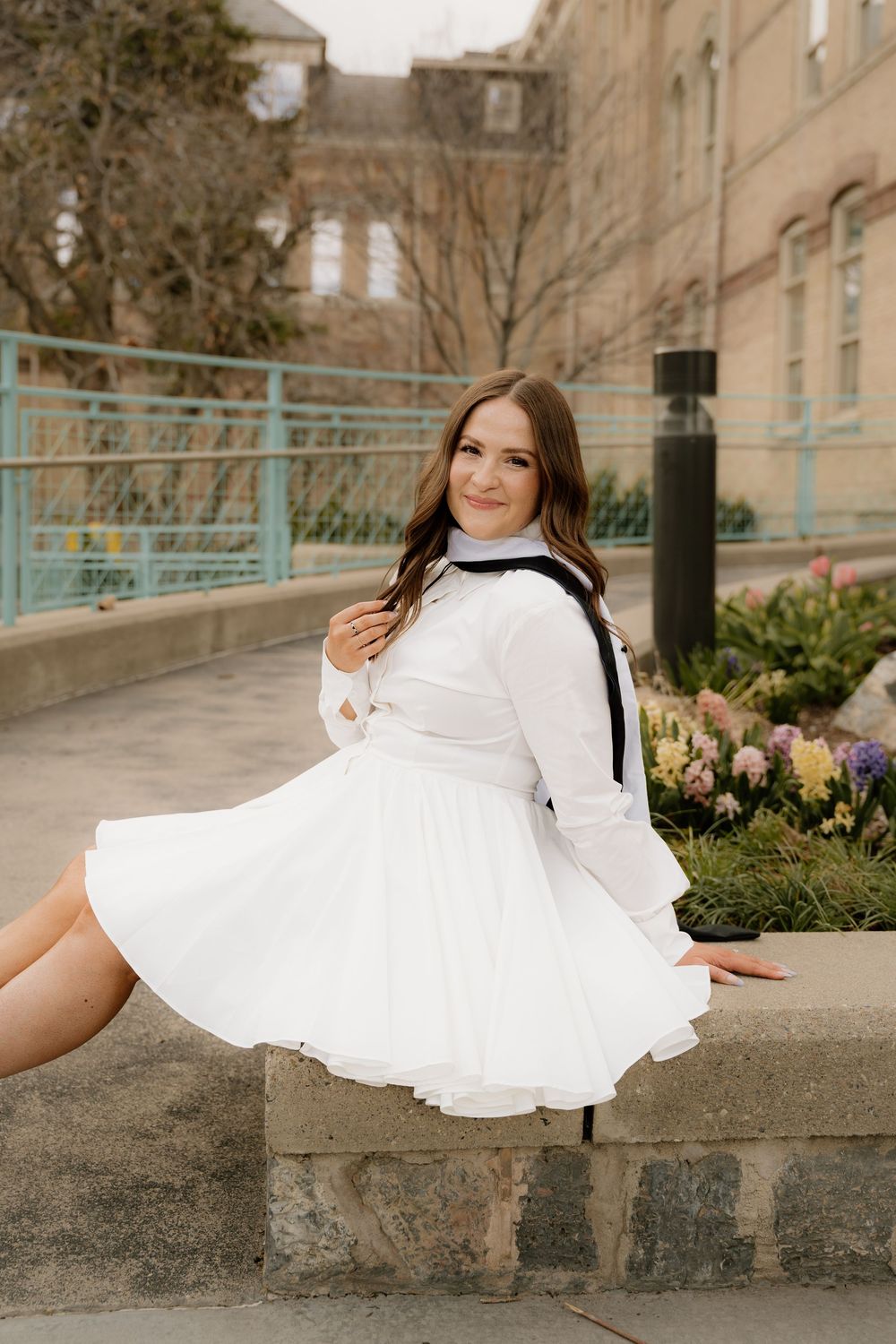 Person in flowing white dress and black accessories sits on stone wall with flowers blooming in garden behind.