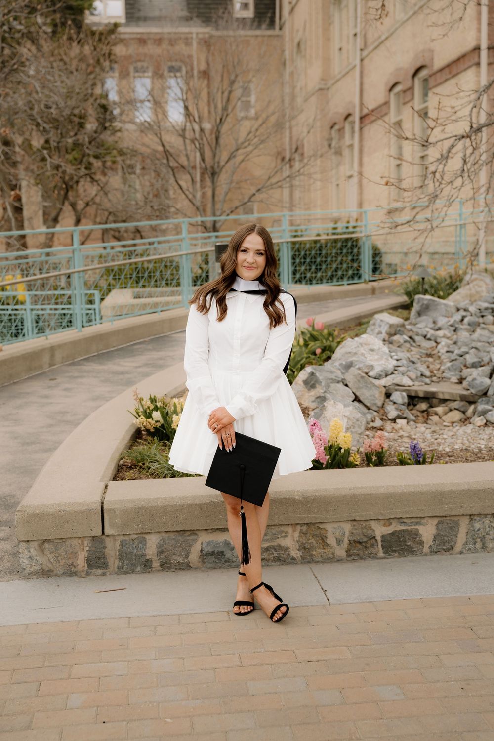 Graduate in white dress poses on concrete steps with retaining wall and campus building in background.