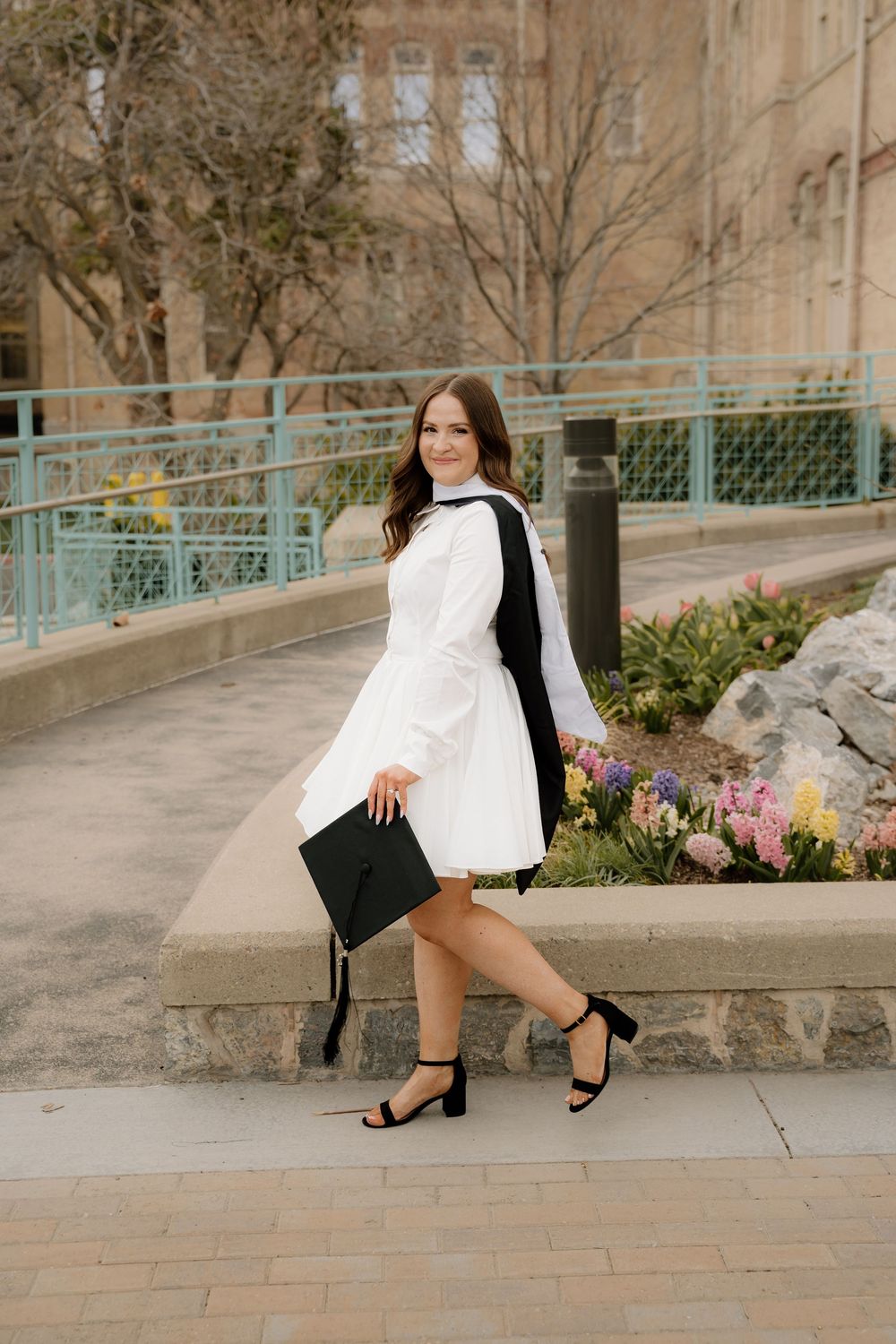 Graduate in white dress holding cap stands near turquoise railing with spring flowers in background.
