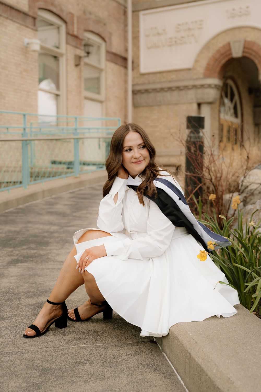 A person in a flowing white dress reclines on concrete steps near blooming daffodils in a spring outdoor setting.
