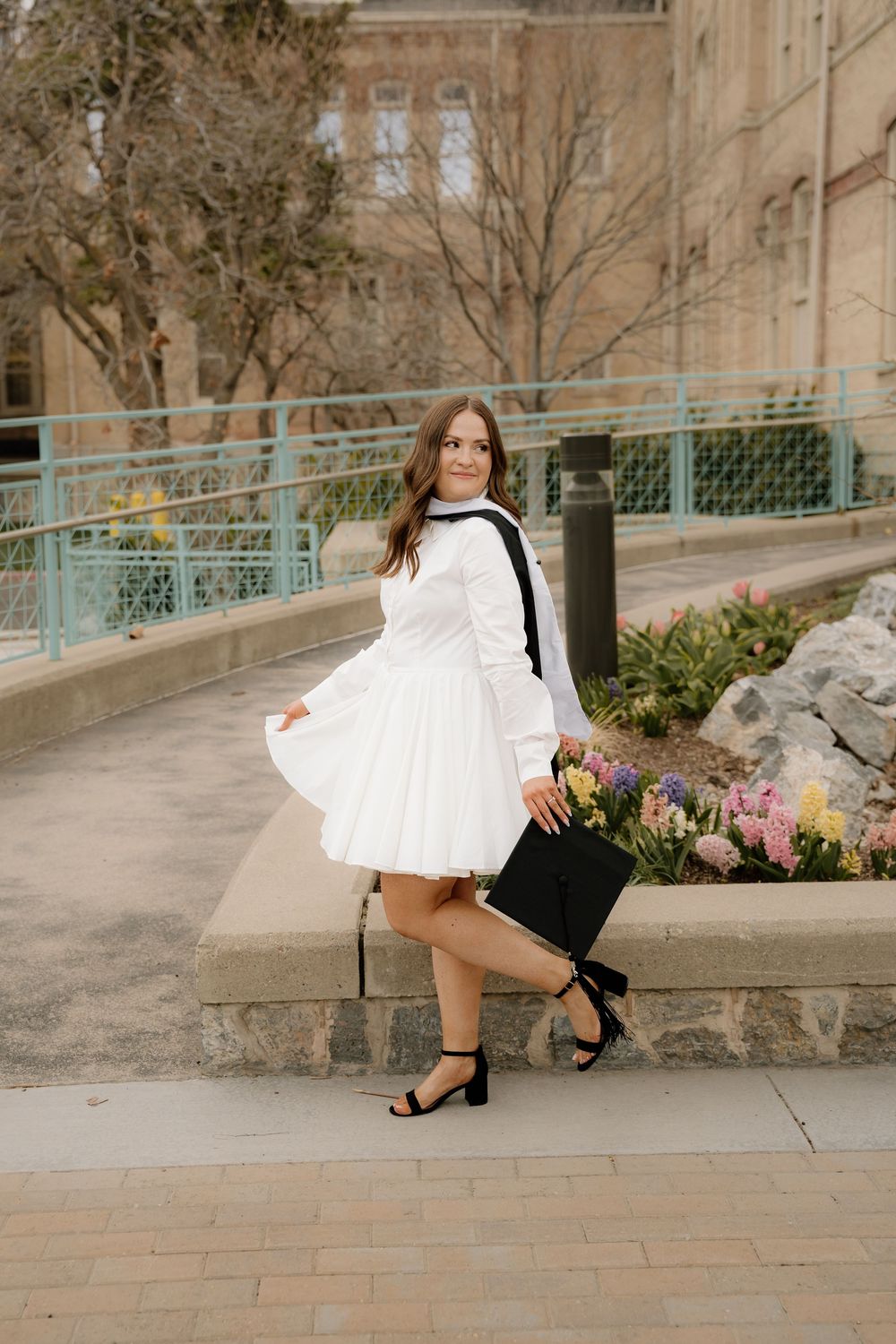 Graduate in flowy white dress poses on campus path near blooming flowers and stone building.