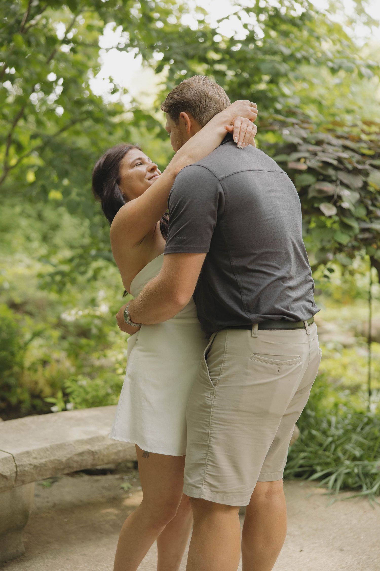 A couple embrace in a romantic pose outdoors surrounded by lush greenery and garden scenery.
