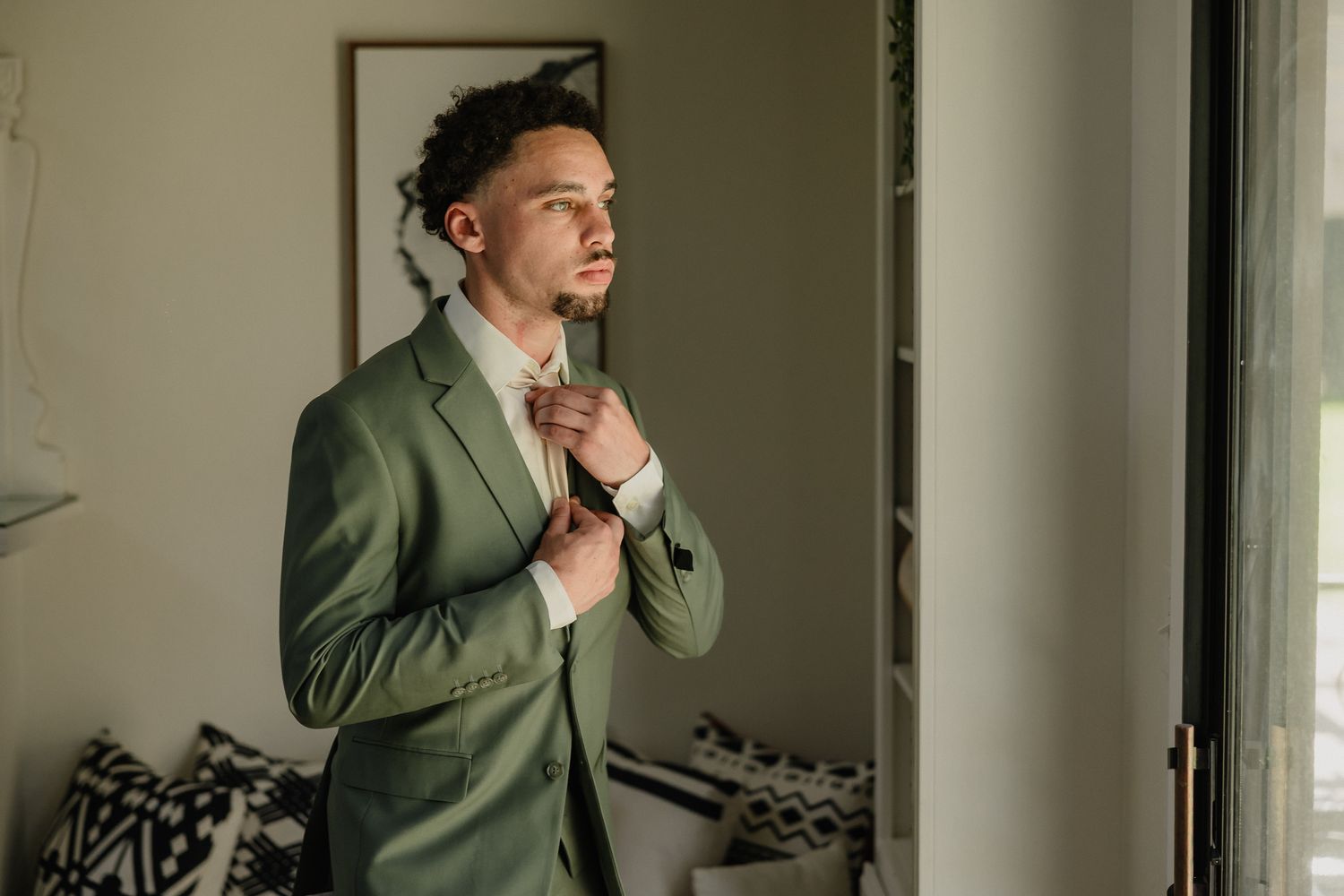 A person in an olive green suit adjusts their bow tie while getting ready.