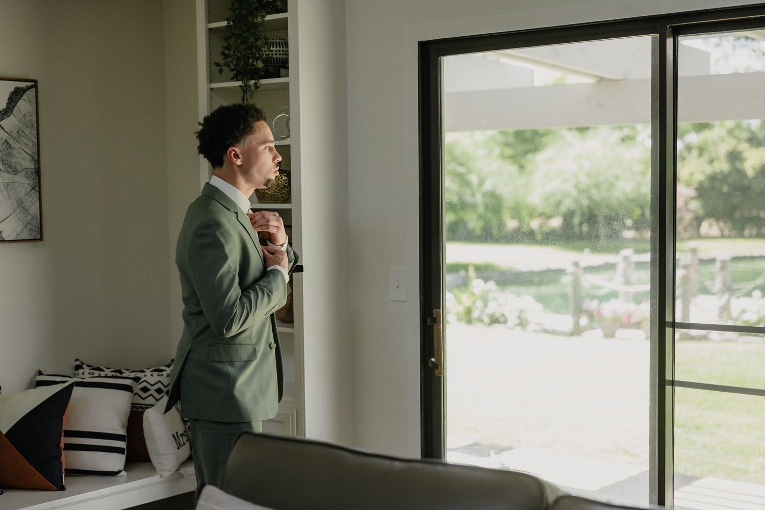 Professional in a grey suit jacket stands at a large office window with natural light streaming in.
