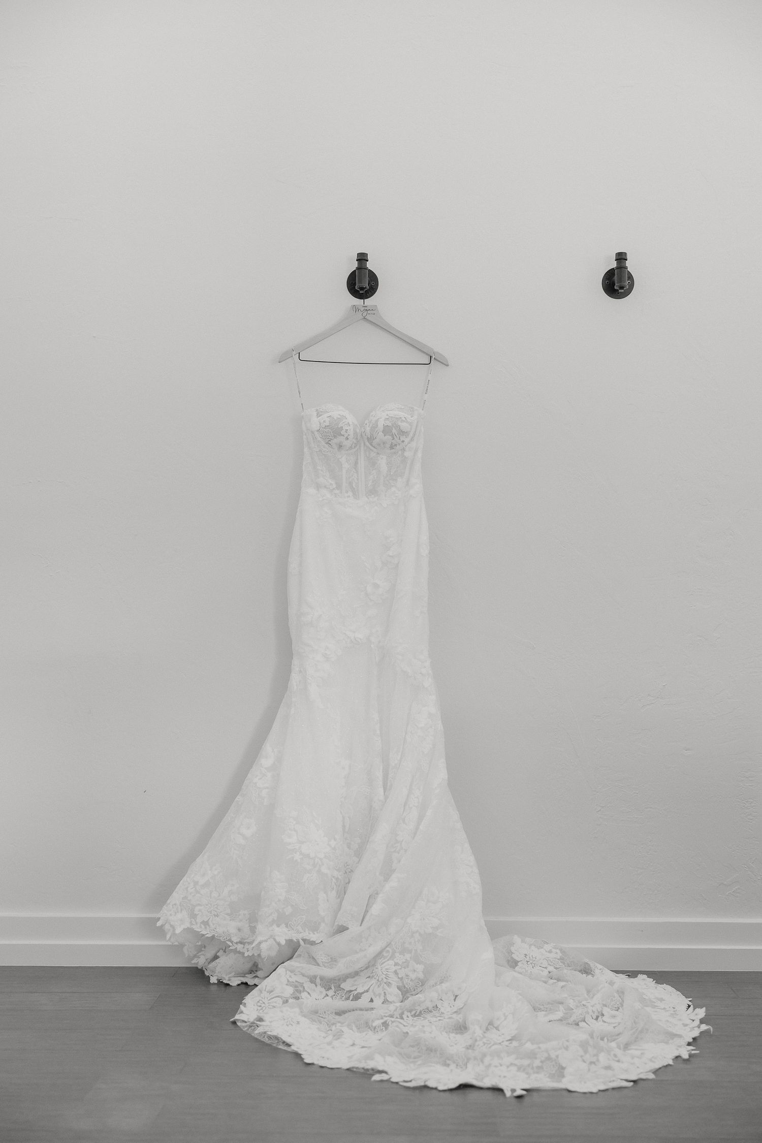 A white lace wedding dress with a long train hangs elegantly against a neutral wall.