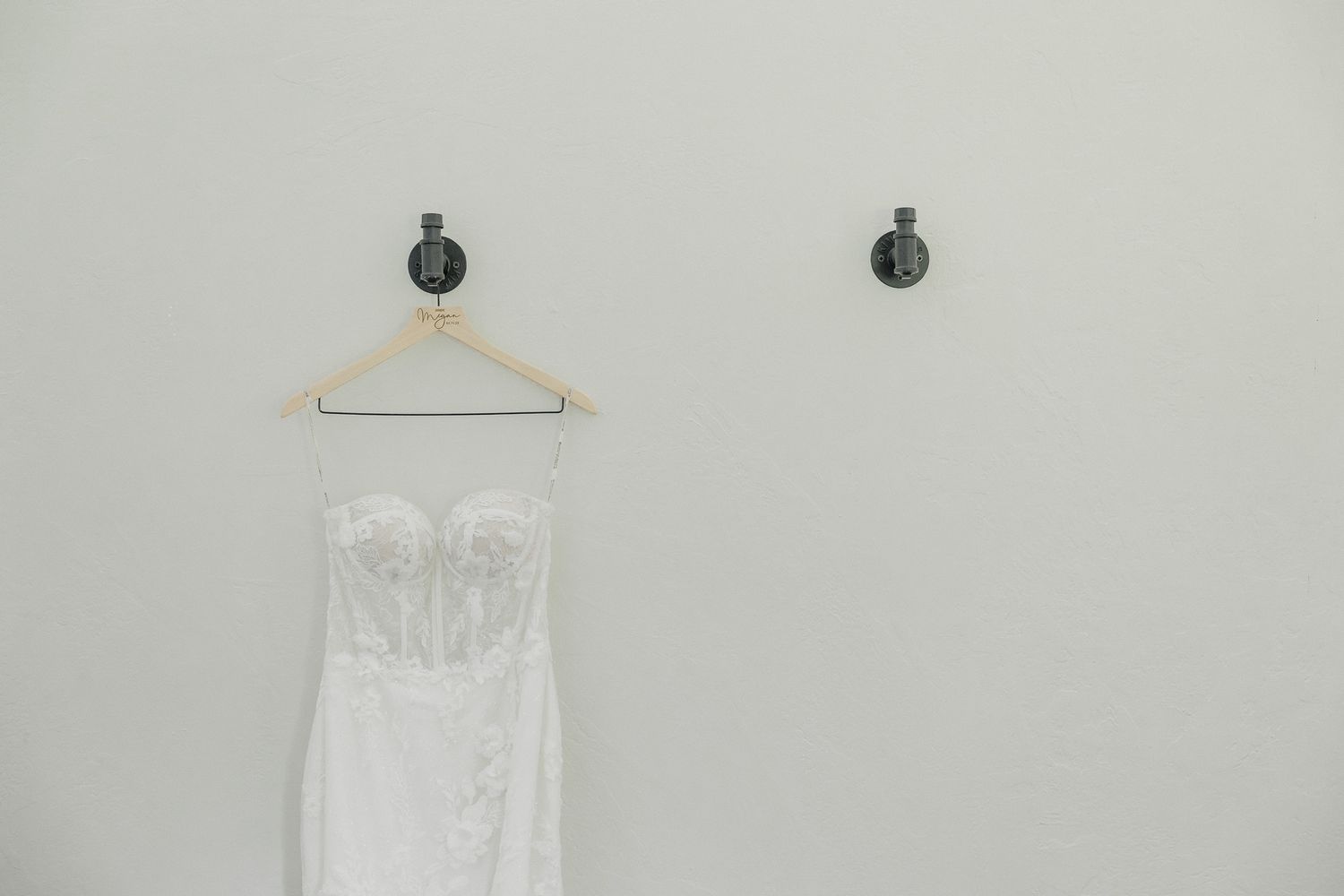 A white strapless wedding dress hangs on a wooden hanger against a light gray wall with two wall sconces.