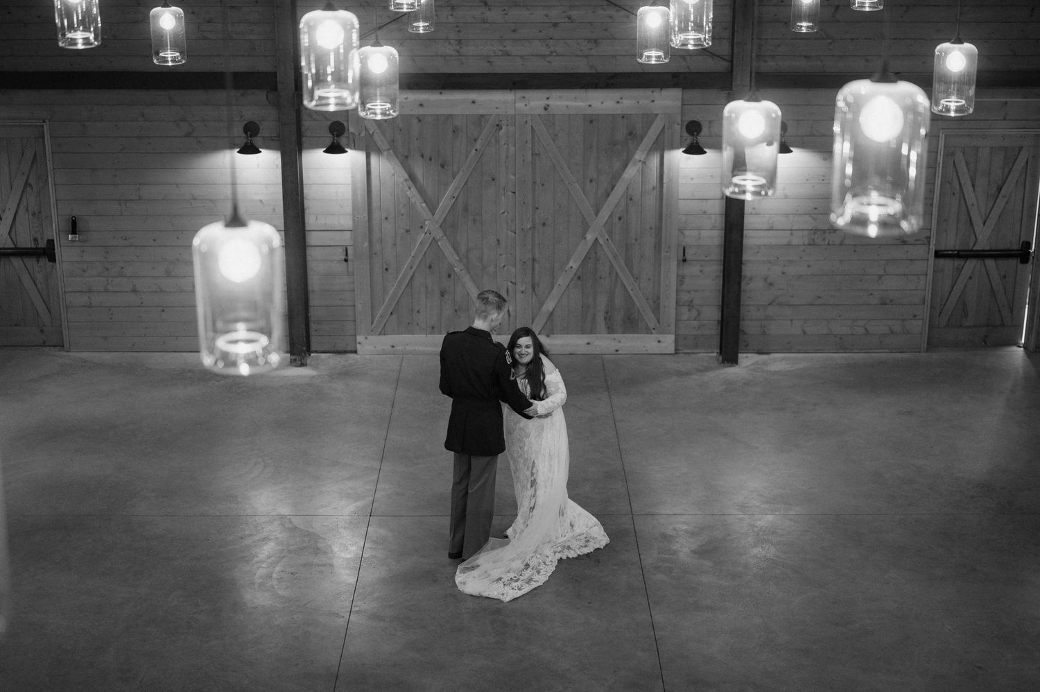 A black and white photograph of a couple dancing in a rustic barn venue with illuminated glass pendant lights overhead.