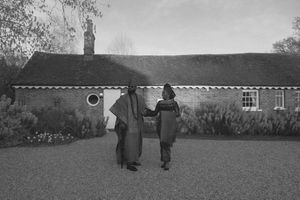 Two figures stand on a lawn in front of a single-story rural cottage with a thatched roof in black and white.