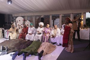 A group in period costumes lies on the floor and sits in chairs during what appears to be a theatrical performance.