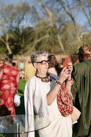 Person in white outfit and glasses taking a photo with mobile phone at outdoor gathering.