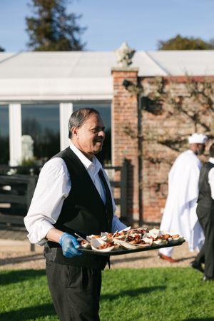 A server in formal attire carries a platter of appetizers at an outdoor catering event.