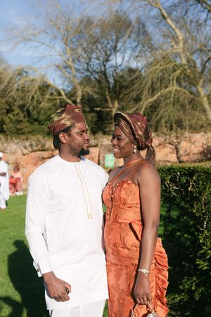 A couple in traditional West African attire, with the woman in orange and the man in white, stand together at an outdoor event.