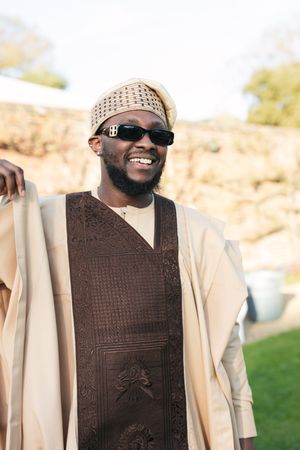 A person wearing sunglasses and traditional African attire smiles during an outdoor celebration.