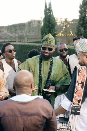 A group dressed in traditional African attire gathered outdoors with cypress trees and string lights in the background.