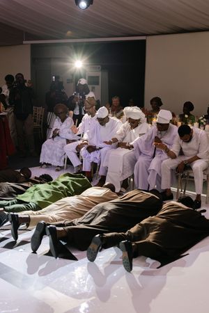 People dressed in all white clothing sit in chairs while others lie down during what appears to be a performance or ceremony.