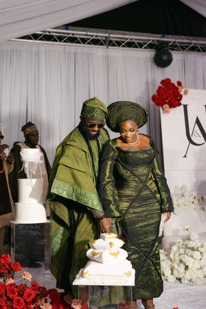 A couple in traditional Nigerian attire cuts their elegant white wedding cake adorned with gold details and red floral decor.