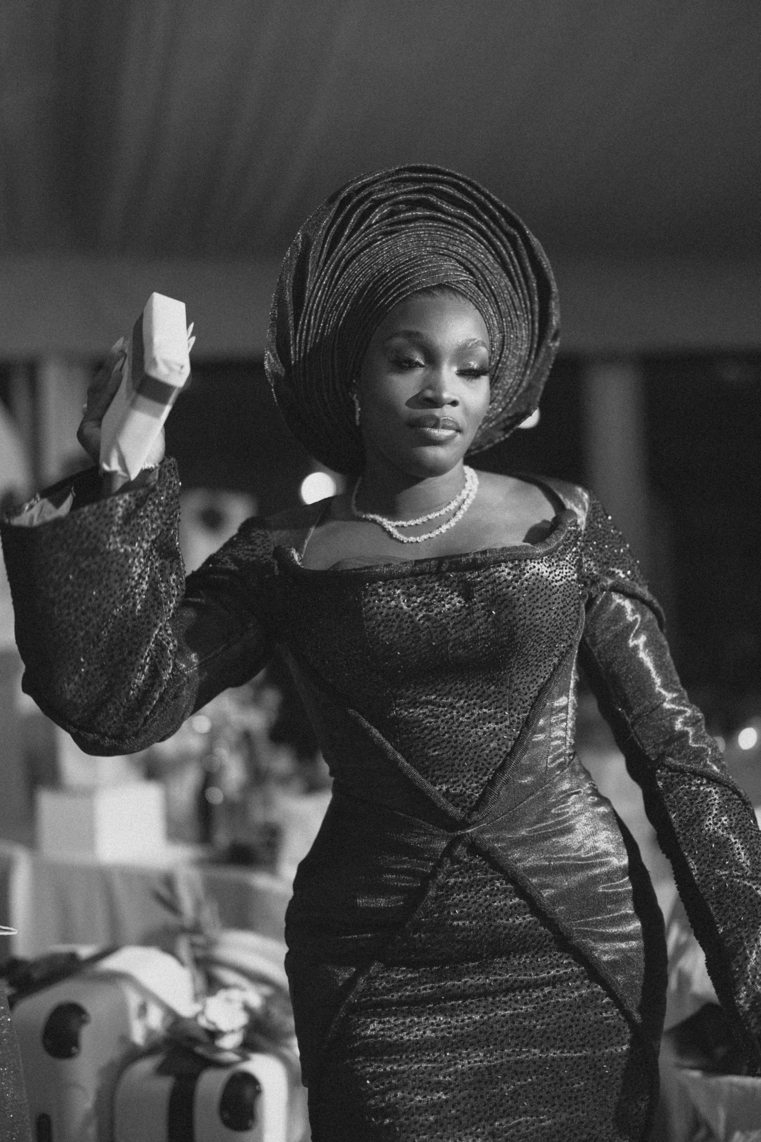 Black and white photo of an elegant woman in a glamorous gown and traditional Nigerian gele head wrap at a formal event.
