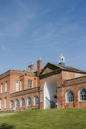 Historic red brick mansion with arched windows and white bell tower sits on grassy lawn under bright blue sky.