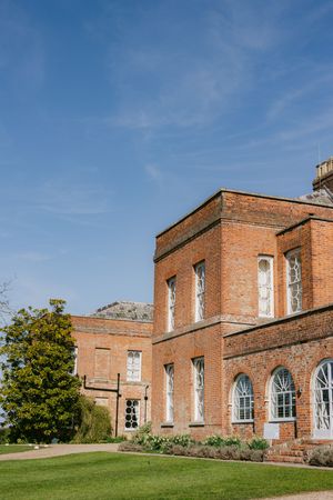 A grand brick Georgian mansion with arched windows and manicured gardens set against a bright blue sky.