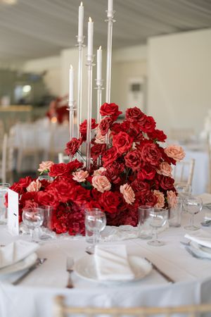 Elegant wedding table centerpiece with tall white candles and lush red and pink roses on white tablecloth.