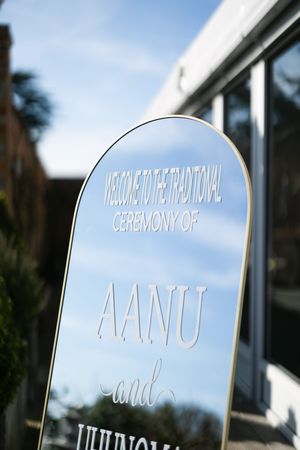 White wedding ceremony sign with reflective mirror finish showing engraved text against a blurred outdoor backdrop.