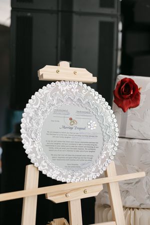 A circular marriage proposal sign displayed on a wooden easel stand with a red rose decoration beside it.