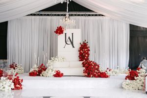 Elegant white wedding cake adorned with cascading red flowers on display against white draping at reception venue.