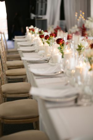 Elegant wedding reception table setting with white linens, candles and red rose centerpieces in a softly lit room.