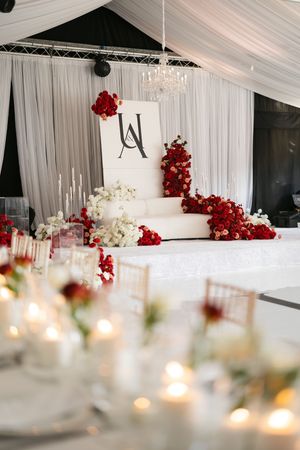 White tiered wedding cake decorated with cascading red flowers and monogram displayed against draped white fabric backdrop.