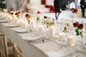 An elegant wedding table setting with white linens, glowing candles, and red rose centerpieces in glass vases.