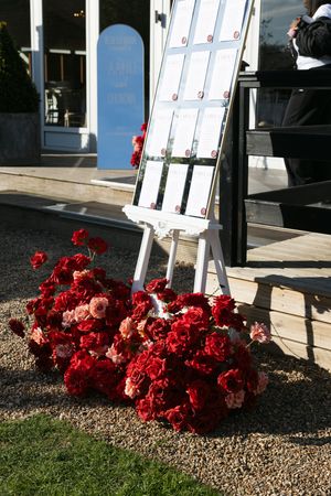 A white seating chart display stand with red floral arrangements at its base outdoors near a venue entrance.