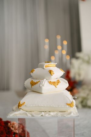 A white wedding ring pillow decorated with gold embroidered details and stacked in a tiered arrangement.